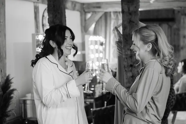 A bride and her bridesmaid clinking champagne glasses at the lit vanity mirror