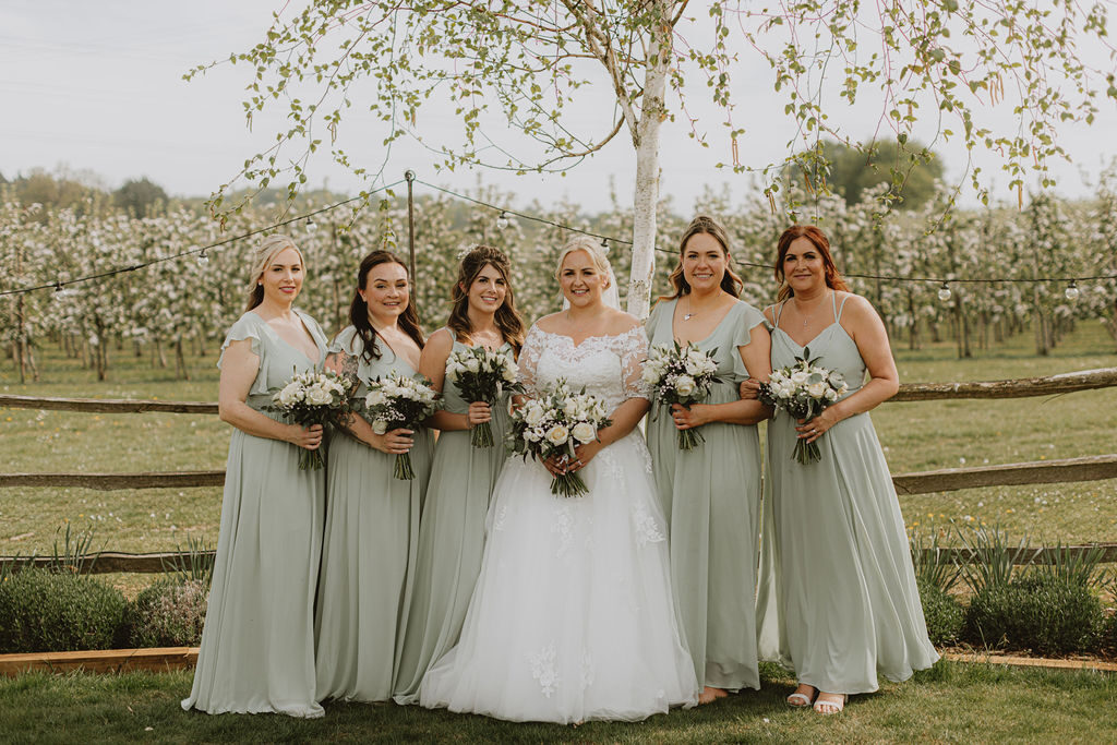 A bride and her bridesmaids in sage green in front of the blossoming orchards at The BarnYard