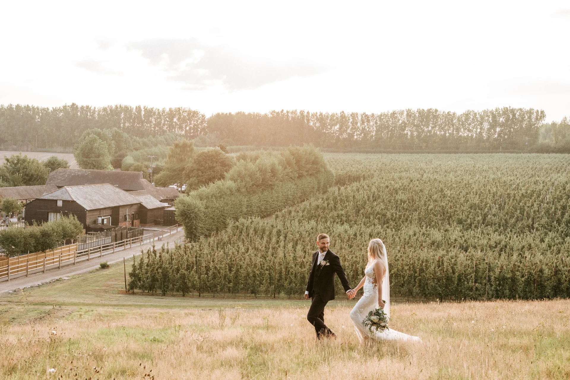 A couple walking hand in hand through the meadow above The BarnYard at golden hour, orchards stretching out below