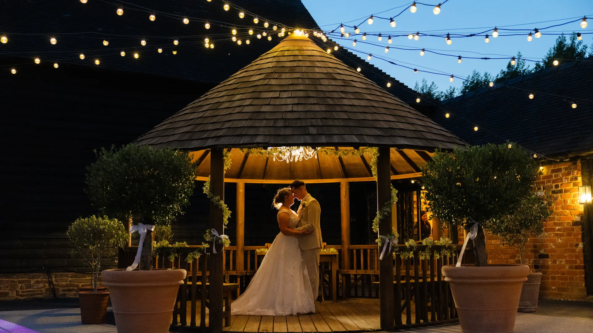 A couple beneath the Courtyard gazebo at twilight, festoon lights strung overhead at The BarnYard, Kent